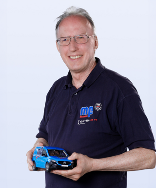 Ein Mann hält ein Modellauto in der Hand Ein älterer Mann mit Brille lächelt, während er ein blaues Modellauto in der Hand hält, das ein Mercedes-Benz darstellt.