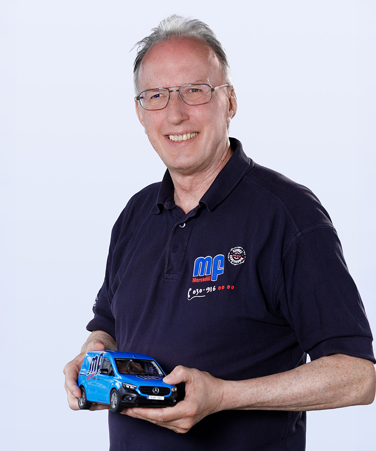 Ein älterer Mann mit Brille lächelt, während er ein blaues Modellauto in der Hand hält, das ein Mercedes-Benz darstellt.