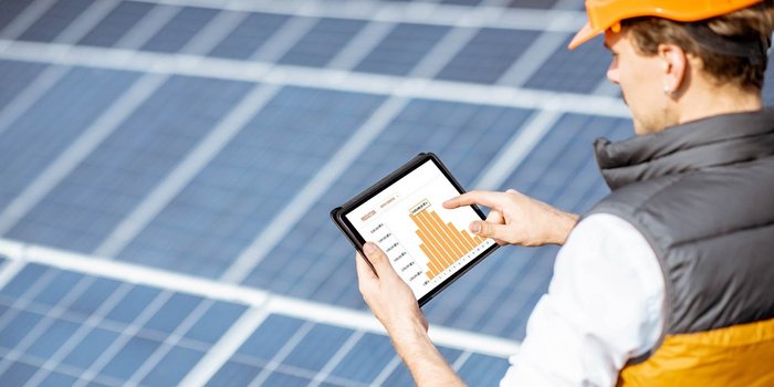 Der Mann mit einem grauen Weste und orangefarbenem Helm überprüft Diagramme auf einem Tablet vor einer Solaranlage.