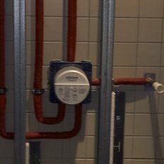 Eine enge Dusche mit grauen Fliesen, in der rote Rohre und ein Wasserzähler sichtbar sind.