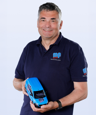 Ein lächelnder Mann in einem blauen Poloshirt hält ein Modell eines blauen Fahrzeugs in seinen Händen.