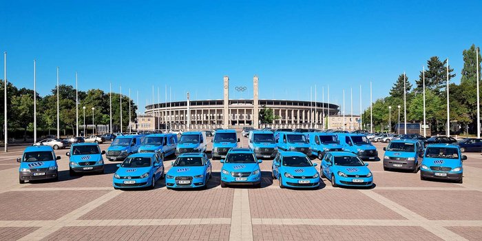 Verschiedene blaue Autos und Vans sind auf einem Platz angeordnet, mit einem großen Stadion im Hintergrund.