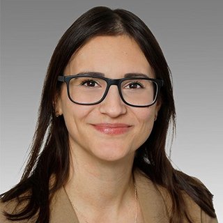 Professionelles Porträt einer lächelnden Frau mit Brille Eine Frau mit langen, dunklen Haaren und Brille lächelt freundlich in die Kamera, vor einem grauen Hintergrund.
