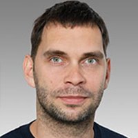 Ein Mann mit kurzen, dunklen Haaren und einem leichten Bart blickt direkt in die Kamera, mit einem neutralen Gesichtsausdruck.