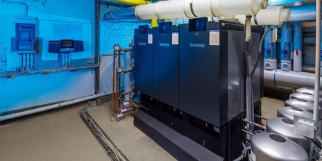 Die Heizungsanlage zeigt drei große Kessel mit digitalen Anzeigen, während Rohre und Ventile im Hintergrund sichtbar sind.
