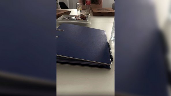 Ein blauer Ordner liegt auf einem Tisch, umgeben von Pizzakartons und Snacks, während eine Person im Hintergrund sitzt.