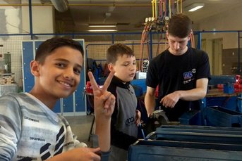 In einer Werkstatt stehen drei Kinder, einer lächelt und macht ein Zeichen, während die anderen konzentriert arbeiten.