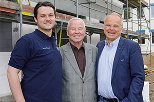 Drei Männer stehen lächelnd vor einem Baugerüst, das auf einer Baustelle im Hintergrund sichtbar ist.