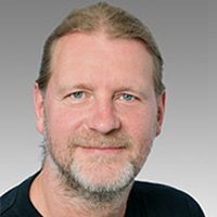 Der Mann hat blaue Augen und trägt ein schwarzes T-Shirt, während er in die Kamera schaut und entspannt wirkt.