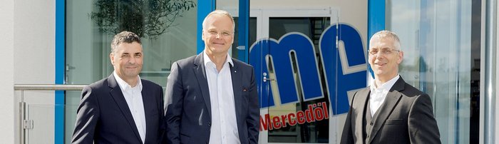 Professionelles Teamfoto vor der Mercedöl-Firma im Freien Drei Männer in Anzügen stehen vor einem modernen Gebäude mit blauen Akzenten und einem großen Logo im Hintergrund.