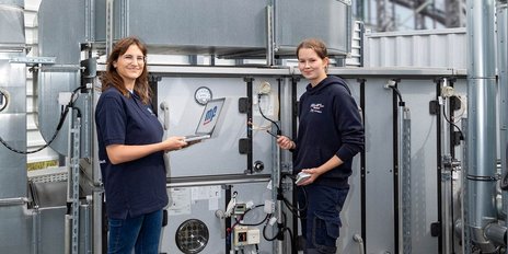 Zwei Frauen stehen vor einer technischen Anlage, eine hält einen Laptop, die andere ein Messgerät, beide lächeln.