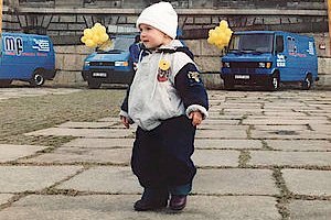 Ein kleines Kind in warmer Kleidung steht auf einem gepflasterten Platz, umgeben von blauen Lieferwagen und gelben Ballons.