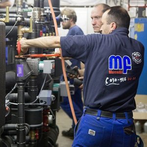 In einer technischen Umgebung repariert ein Handwerker mit einem Werkzeug eine Maschine, während andere Mitarbeiter helfen.