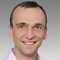 Professionelles Porträt eines freundlichen Mannes Der Mann hat eine positive Ausstrahlung, mit einem breiten Lächeln und klaren, blauen Augen vor einem grauen Hintergrund.
