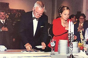 Ein elegant gekleideter Mann schneidet eine Torte, während eine Frau in einem roten Kleid Geschirr auf einem Tisch anordnet.