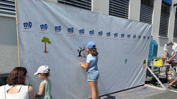 Eine Gruppe von Kindern malt an einer Wand, während Erwachsene Materialien vorbereiten und die Szene beobachten.