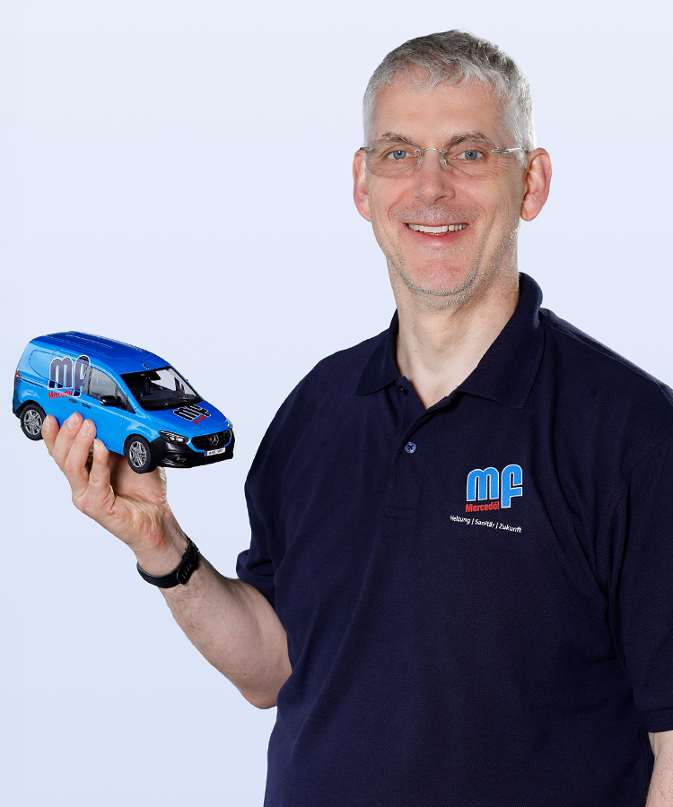Ein lächelnder Mann hält ein Modell eines blauen Lieferwagens in der Hand, während er in einem dunklen Poloshirt steht.