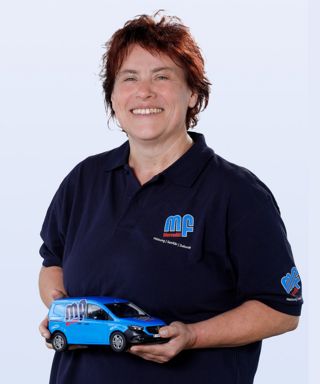 Frau mit Modellauto in blauer Arbeitskleidung lächelt Eine lächelnde Frau mit kurzen, roten Haaren hält ein Modell eines blauen Mercedes-Vans in der Hand.