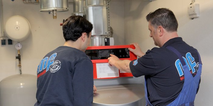 Zwei Männer stehen in einem technischen Raum und überprüfen gemeinsam ein rotes Bedienfeld an einer Maschine.