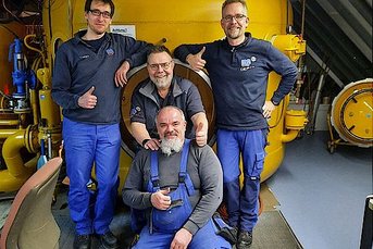 Vier Männer posieren lächelnd vor einem großen gelben Gerät in einem technischen Raum, während einer Daumen hoch zeigt.