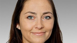 Eine Frau mit langen, dunklen Haaren und strahlend blauen Augen lächelt freundlich in die Kamera vor einem grauen Hintergrund.