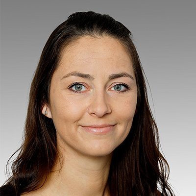 Eine Frau mit langen, dunklen Haaren und strahlend blauen Augen lächelt freundlich in die Kamera vor einem grauen Hintergrund.