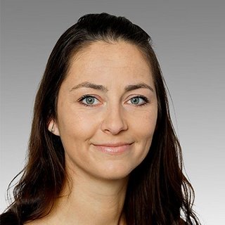 Porträt einer Frau mit strahlendem Lächeln und langen Haaren Eine Frau mit langen, dunklen Haaren und strahlend blauen Augen lächelt freundlich in die Kamera vor einem grauen Hintergrund.