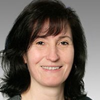 Porträt einer freundlichen Frau mit natürlichem Ausdruck Eine Frau mit dunklen, lockigen Haaren lächelt freundlich in die Kamera, mit einem neutralen grauen Hintergrund.