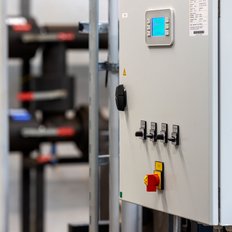 Ein Steuerungspanel mit einem blauen Bildschirm und mehreren Tasten steht in einer industriellen Umgebung mit Rohren im Hintergrund.