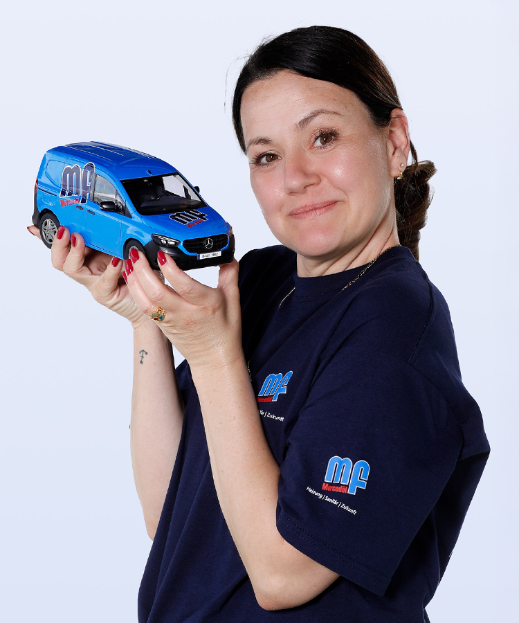 Eine Frau hält ein Modell eines blauen Mercedes-Vans in ihren Händen und lächelt dabei freundlich in die Kamera.