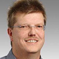Professionelles Porträt eines lächelnden Mannes mit Brille Der Mann zeigt ein freundliches Lächeln, während er in einem hellen, professionellen Umfeld abgebildet ist.
