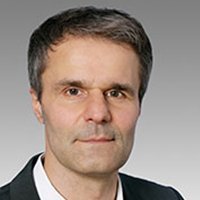 Ein Mann mit kurzen, grauen Haaren und einem neutralen Gesichtsausdruck trägt einen schwarzen Anzug und ein weißes Hemd.