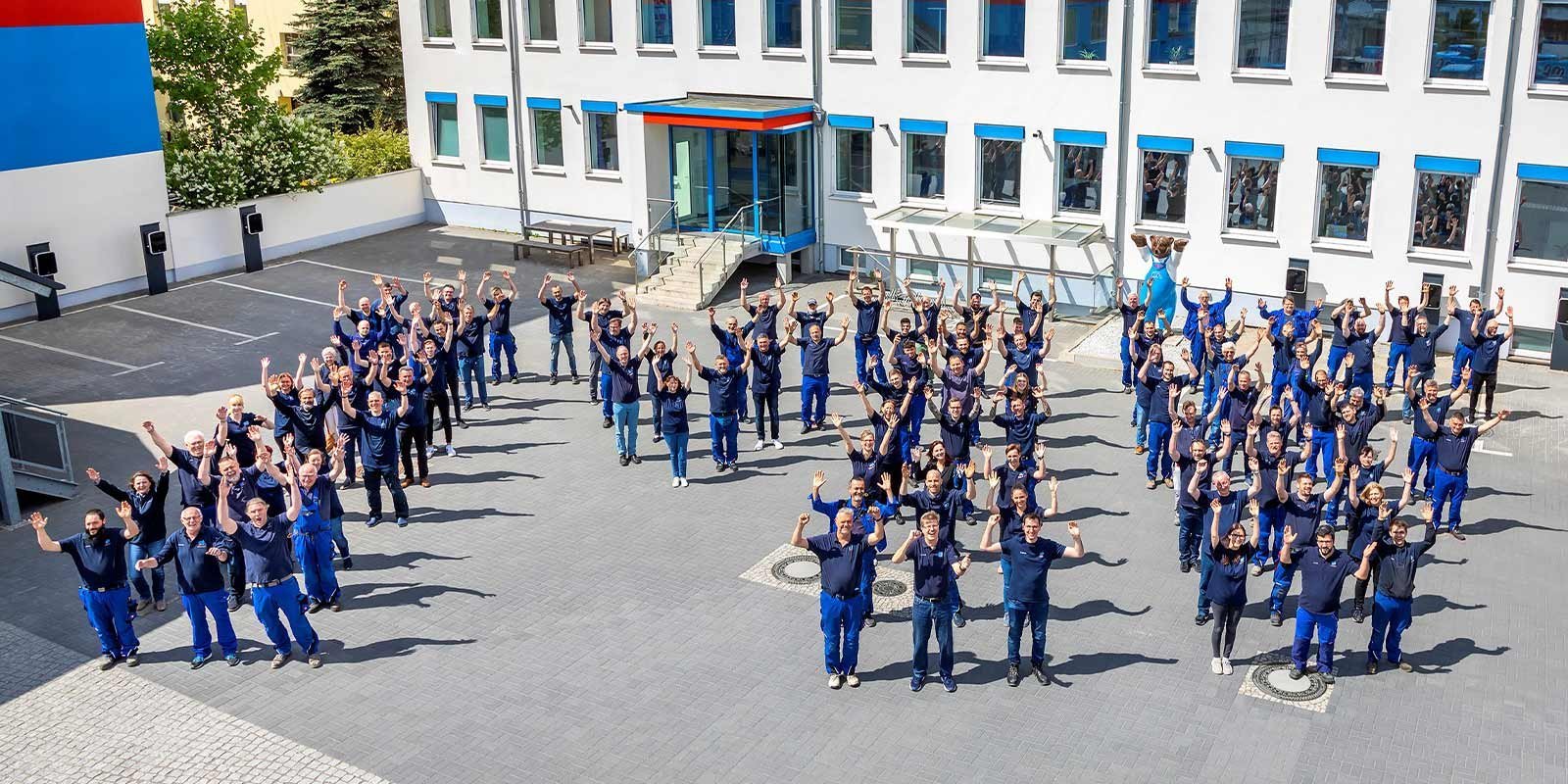 Eine große Gruppe von Menschen in blauen Arbeitsanzügen versammelt sich im Freien und hebt die Hände in die Luft.