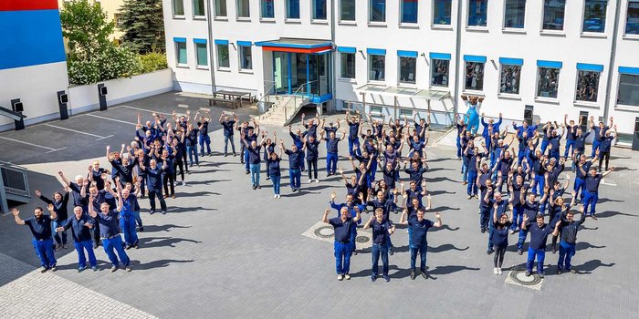 Eine große Gruppe von Menschen in blauen Arbeitsanzügen versammelt sich im Freien und hebt die Hände in die Luft.