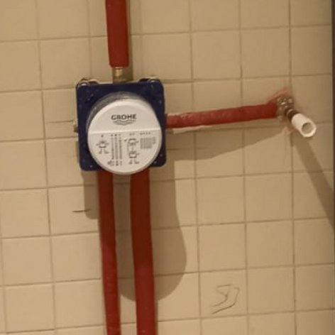 Eine Wand mit weißen Fliesen, an der ein Wasserzähler und rote Rohre für die Wasserleitung montiert sind.