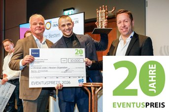Ein Gewinner hält stolz einen großen Scheck über 10.000 Euro und eine Auszeichnung in der Hand, umgeben von jubelnden Anwesenden.