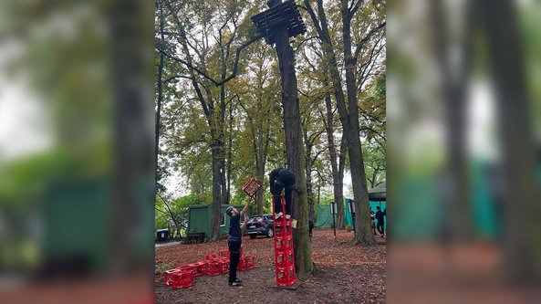 Zwei Personen stehen in einem Wald, während eine Person auf einem Turm aus roten Kisten balanciert, um eine Plattform zu erreichen.