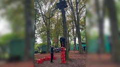 Zwei Personen stehen in einem Wald, während eine Person auf einem Turm aus roten Kisten balanciert, um eine Plattform zu erreichen.