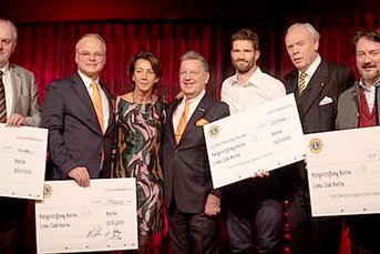 Die Gruppe von sechs Menschen, darunter eine Frau in einem bunten Kleid, präsentiert stolz große Schecks bei einer Veranstaltung.