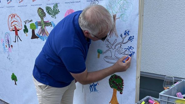 Ein Mann mit blonden Haaren zeichnet mit einem blauen Stift auf eine große, bemalte Wand voller bunter Zeichnungen.