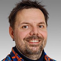 Ein lächelnder Mann mit Bart trägt ein kariertes Hemd und schaut direkt in die Kamera, Hintergrund ist grau.