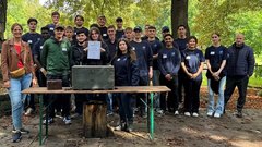 Eine Gruppe von jungen Erwachsenen steht lächelnd vor einem Tisch mit einer Kiste und einem Zertifikat in einem Park.