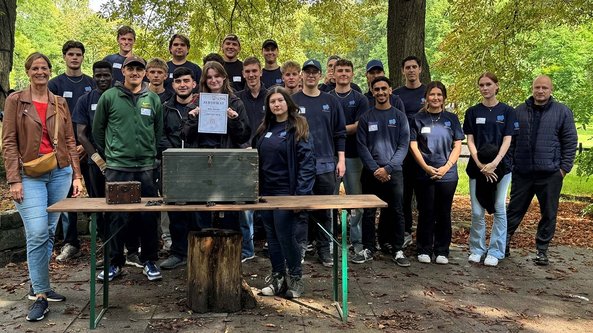 Eine Gruppe von jungen Erwachsenen steht lächelnd vor einem Tisch mit einer Kiste und einem Zertifikat in einem Park.