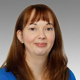 Professionelles Porträt einer lächelnden Frau mit braunen Haaren Eine Frau mit langen, braunen Haaren und einem freundlichen Lächeln trägt ein blaues Oberteil vor einem grauen Hintergrund.
