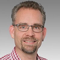Ein Mann mit Brille und einem freundlichen Lächeln trägt ein kariertes Hemd und hat einen gepflegten Bart.