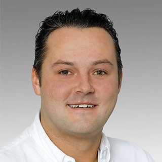 Professionelles Porträt eines lächelnden Mannes in Weiß Ein Mann mit dunklen, lockigen Haaren lächelt freundlich in die Kamera, trägt ein weißes Hemd und hat einen neutralen Hintergrund.