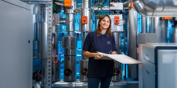 In einem industriellen Umfeld steht eine Frau mit Brille, die ein Blatt Papier hält, während sie von Maschinen umgeben ist.