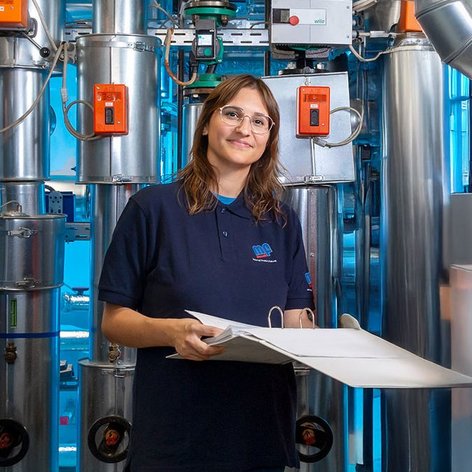 In einem industriellen Umfeld steht eine Frau mit Brille, die ein Blatt Papier hält, während sie von Maschinen umgeben ist.