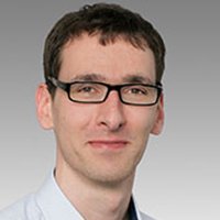 Professionelles Porträt eines Mannes mit Brille Ein Mann mit Brille und kurzem Haar lächelt freundlich in die Kamera, vor einem grauen Hintergrund.