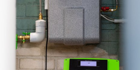 An einer Wand montierte grüne Steuerungseinheit mit digitalem Display und verschiedenen Anschlüssen für Wasserleitungen.